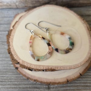 May include: A pair of teardrop-shaped earrings with a colorful, marbled pattern. The earrings have silver-colored hooks and are displayed on a wooden slice. The earrings feature shades of beige, green, and brown.