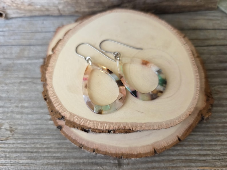 May include: A pair of teardrop-shaped earrings with a colorful, marbled pattern. The earrings have silver-colored hooks and are displayed on a wooden slice. The earrings feature shades of beige, green, and brown.
