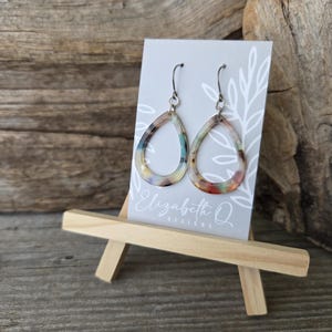 May include: A pair of teardrop-shaped earrings with a colorful, marbled pattern. The earrings are displayed on a small easel with a card that reads "Elizabeth Q Designs". The earrings have silver-colored hooks.