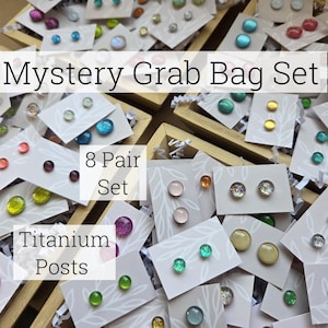 May include: Mystery Grab Bag Set of 8 pairs of earrings with titanium posts. The earrings are displayed on cards with white backgrounds and green leaf designs. The earrings are made of glass and come in a variety of colors and patterns.