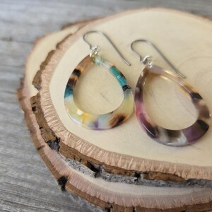 May include: A pair of teardrop-shaped earrings with silver hooks. The earrings have a marbled pattern with colors including teal, yellow, brown, and purple. The earrings are displayed on a wooden slice.