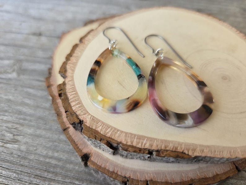 May include: A pair of teardrop-shaped earrings with silver hooks. The earrings have a marbled pattern with colors including teal, yellow, brown, and purple. The earrings are displayed on a wooden slice.