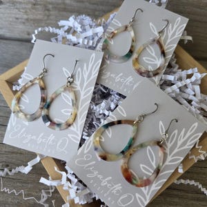 May include: A wooden box filled with white paper shreds holds several pairs of teardrop earrings. The earrings have a marbled pattern in shades of brown, green, and cream. Each pair is attached to a gray card with the text "Elizabeth Q Designs".