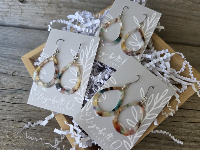 May include: A wooden box filled with white paper shreds holds several pairs of teardrop earrings. The earrings have a marbled pattern in shades of brown, green, and cream. Each pair is attached to a gray card with the text "Elizabeth Q Designs".