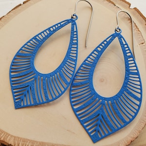 May include: A pair of vibrant blue teardrop earrings with a textured surface and intricate cut-out design. The earrings have silver-colored ear wires and are displayed on a light wood surface.