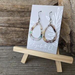 May include: A pair of teardrop-shaped earrings with a marbled pattern in shades of brown, beige, and green. The earrings are displayed on a small easel with a gray card featuring the text "Elizabeth Q Designs".