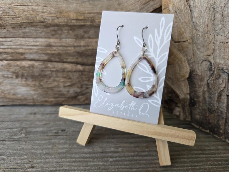 May include: A pair of teardrop-shaped earrings with a marbled pattern in shades of brown, beige, and green. The earrings are displayed on a small easel with a gray card featuring the text "Elizabeth Q Designs".