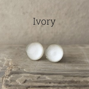 May include: Two ivory-colored, round stud earrings. The earrings are set against a neutral background and rest on a textured, wooden surface. The word "Ivory" is displayed above the earrings.