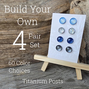 May include: A display of four pairs of stud earrings in various shades of blue, silver, and gray, presented on a small wooden easel. The card reads "Build Your Own 4 Pair Set" with "60 Color Choices" and "Titanium Posts".