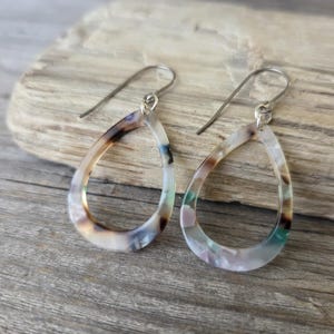 May include: A pair of teardrop earrings with a marbled pattern in shades of brown, white, and teal. The earrings have silver-colored ear wires and are displayed on a piece of driftwood.