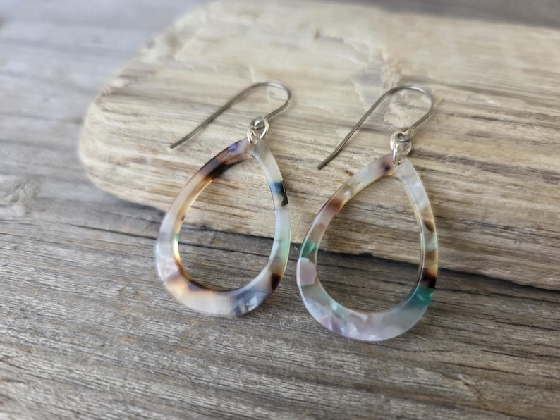 May include: A pair of teardrop earrings with a marbled pattern in shades of brown, white, and teal. The earrings have silver-colored ear wires and are displayed on a piece of driftwood.