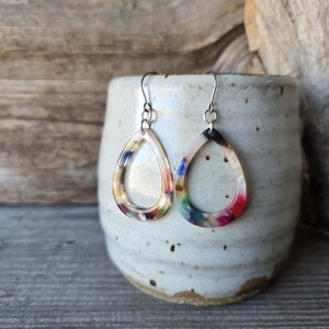May include: A pair of teardrop earrings with a colorful, marbled design. The earrings are suspended from silver-colored hooks and are displayed against a rustic, ceramic container.