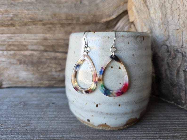 May include: A pair of teardrop earrings with a colorful, marbled design. The earrings are suspended from silver-colored hooks and are displayed against a rustic, ceramic container.