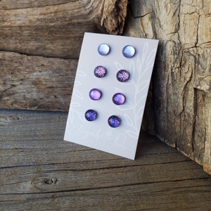 May include: Set of six purple and lavender stud earrings with glitter accents. The earrings are displayed on a white card with the text "Elizabeth Q".
