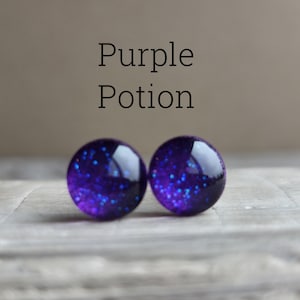 May include: A pair of round stud earrings featuring a deep purple base with embedded blue glitter particles. The text "Purple Potion" is printed in black above the earrings. The earrings have a smooth, glossy, domed surface that reflects light.