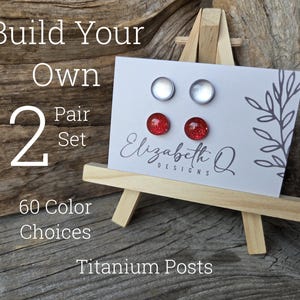 May include: A display of stud earrings on a small wooden easel. The earrings are on a white card with the text "Build Your Own 2 Pair Set" and "60 Color Choices". The earrings include clear and red glitter studs. The card also has the text "Elizabeth Q Designs" and "Titanium Posts".