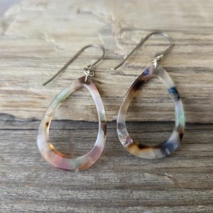 May include: A pair of teardrop-shaped earrings with a marbled pattern in shades of pink, brown, and blue. The earrings have silver-colored hooks and are displayed on a wooden surface.