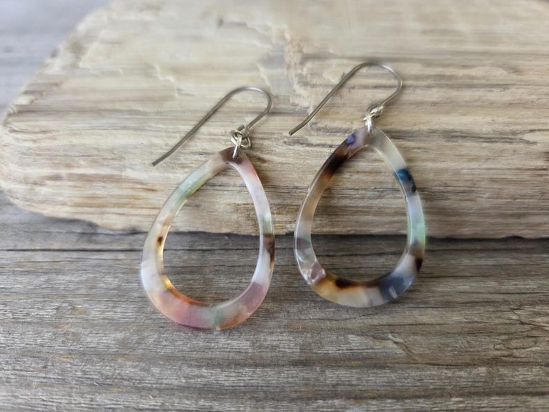 May include: A pair of teardrop-shaped earrings with a marbled pattern in shades of pink, brown, and blue. The earrings have silver-colored hooks and are displayed on a wooden surface.