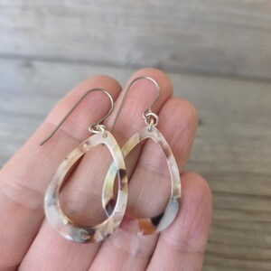 May include: A pair of teardrop-shaped earrings with a marbled pattern in shades of brown, cream, and tan. The earrings have silver-colored ear wires and a hollow center. The earrings are held in a hand.