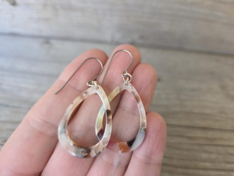 May include: A pair of teardrop-shaped earrings with a marbled pattern in shades of brown, cream, and tan. The earrings have silver-colored ear wires and a hollow center. The earrings are held in a hand.