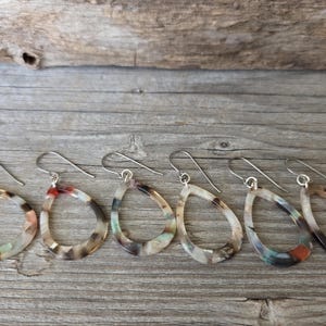 May include: A collection of teardrop-shaped earrings with silver-colored hooks. The earrings have a marbled pattern in shades of brown, beige, and hints of red and green. They are displayed on a weathered wooden surface.