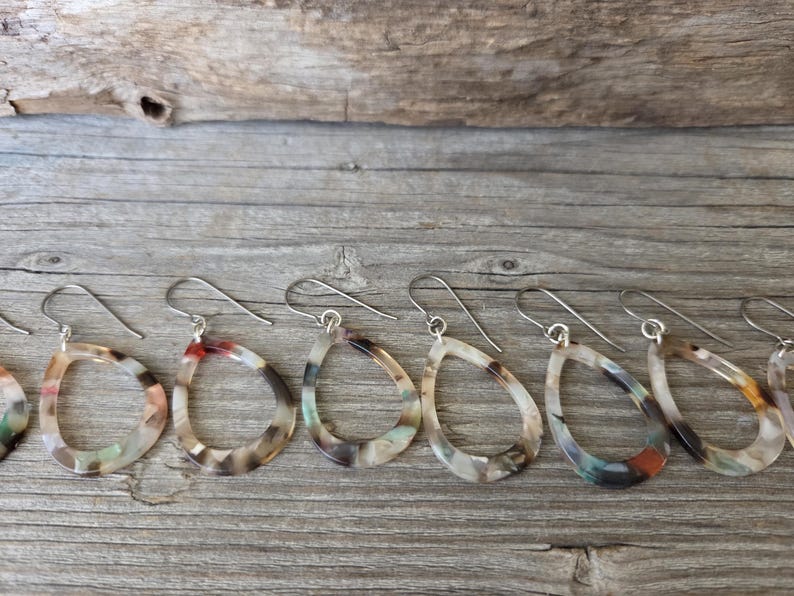 May include: A collection of teardrop-shaped earrings with silver-colored hooks. The earrings have a marbled pattern in shades of brown, beige, and hints of red and green. They are displayed on a weathered wooden surface.