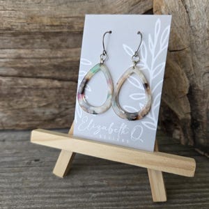 May include: A pair of teardrop-shaped earrings with a marbled pattern in shades of cream, brown, and pastel colors. The earrings are displayed on a small easel with a gray card that has the text "Elizabeth Q Designs".
