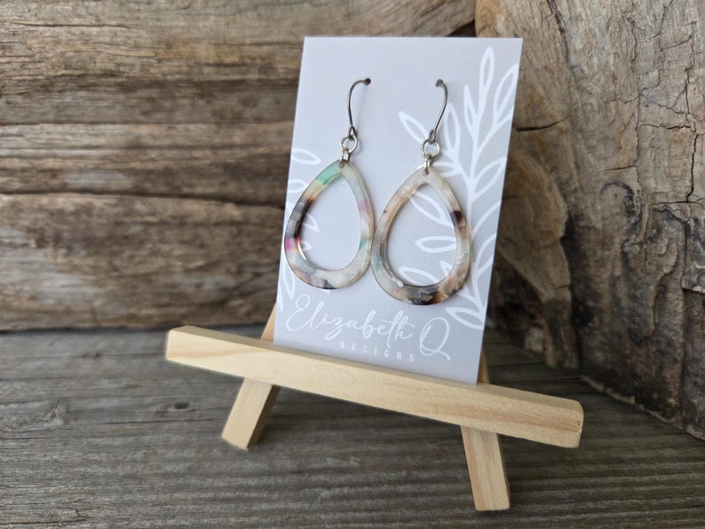 May include: A pair of teardrop-shaped earrings with a marbled pattern in shades of cream, brown, and pastel colors. The earrings are displayed on a small easel with a gray card that has the text "Elizabeth Q Designs".