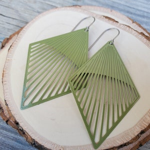 May include: A pair of olive green geometric earrings. Each earring is a diamond shape with a series of parallel lines radiating from the top. The earrings are displayed on a wooden surface.
