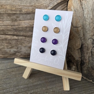 May include: A display of six pairs of stud earrings in various colors: turquoise, tan, purple, and navy blue. The earrings are mounted on a white card with a leaf design and the text "Elizabeth Q Designs". The card is displayed on a small wooden easel.