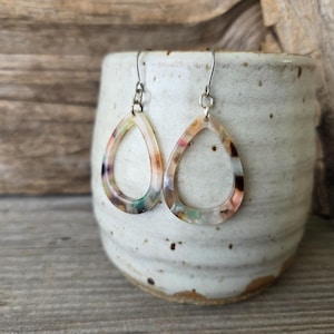 May include: A pair of teardrop-shaped earrings with a colorful, marbled pattern. The earrings are suspended from silver-colored hooks and are displayed in front of a speckled, off-white ceramic mug.