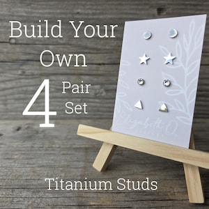 May include: A display of titanium stud earrings on a small wooden easel. The earrings are in various shapes: moons, stars, circles, and triangles. The text reads "Build Your Own 4 Pair Set Titanium Studs."