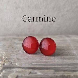 May include: Two carmine-colored, round earrings. The earrings are a vibrant red with a glossy finish. The word "Carmine" is written above the earrings in a black, sans-serif font. The earrings are displayed on a textured, gray surface.