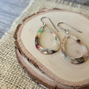 May include: A pair of teardrop-shaped earrings with a colorful, marbled design. The earrings feature a mix of green, pink, and brown hues, suspended from silver-colored ear wires. The earrings are displayed on a wooden slice.