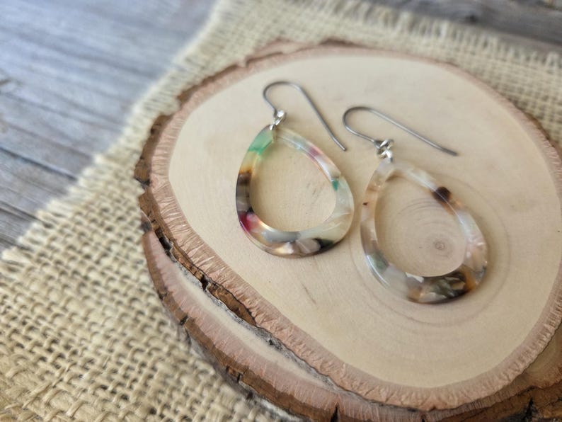 May include: A pair of teardrop-shaped earrings with a colorful, marbled design. The earrings feature a mix of green, pink, and brown hues, suspended from silver-colored ear wires. The earrings are displayed on a wooden slice.