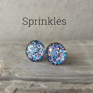 May include: Two round stud earrings with a glittery design. The earrings are covered in a mix of blue, red, and purple glitter, with the word "Sprinkles" in black text above them. The earrings are set against a neutral background.