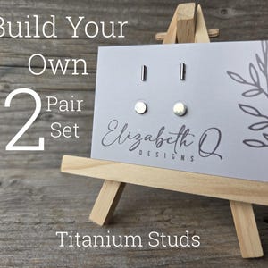 May include: A display of titanium stud earrings on a small wooden easel. The card reads "Build Your Own 2 Pair Set" and "Elizabeth Q Designs". The earrings include two pairs of round studs and two pairs of bar studs.