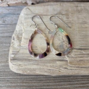 May include: A pair of teardrop-shaped earrings with a marbled pattern in shades of purple, brown, and green. The earrings have silver-colored ear wires and are displayed on a light-colored wooden surface.