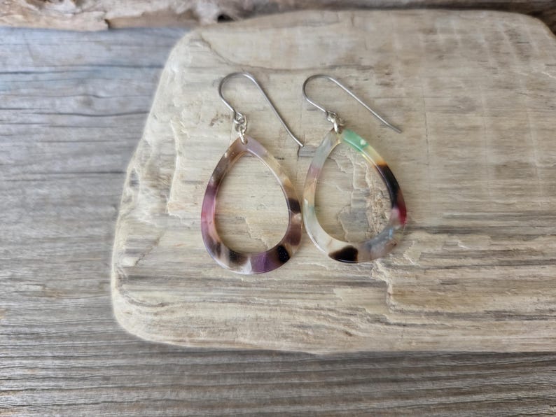 May include: A pair of teardrop-shaped earrings with a marbled pattern in shades of purple, brown, and green. The earrings have silver-colored ear wires and are displayed on a light-colored wooden surface.