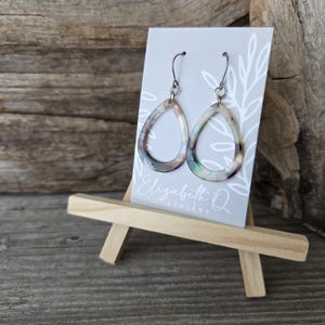 May include: Teardrop-shaped earrings with iridescent accents hang from silver-colored hooks. The earrings are displayed on a small easel, with a gray card featuring the text "Elizabeth Q DESIGNS". The background is a rustic wooden surface.
