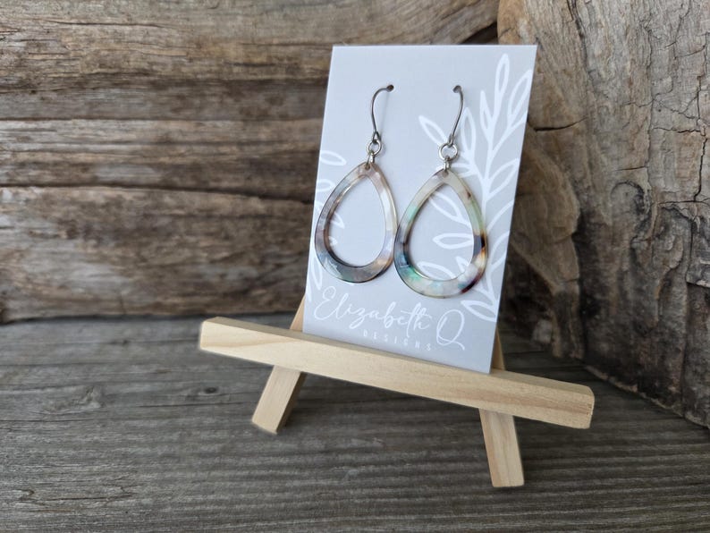 May include: Teardrop-shaped earrings with iridescent accents hang from silver-colored hooks. The earrings are displayed on a small easel, with a gray card featuring the text "Elizabeth Q DESIGNS". The background is a rustic wooden surface.