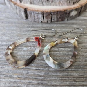 May include: A pair of open teardrop earrings with a marbled pattern in shades of brown, white, and red. The earrings have silver-colored ear wires and are displayed on a wooden surface.
