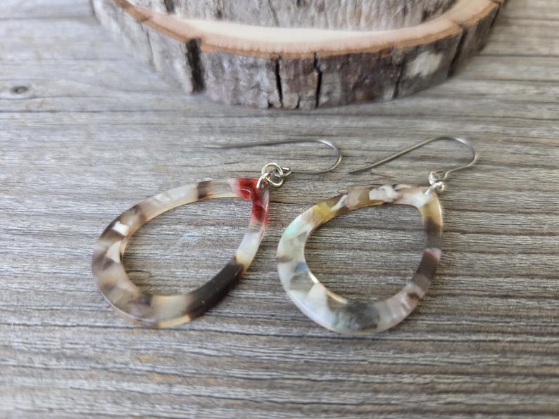 May include: A pair of open teardrop earrings with a marbled pattern in shades of brown, white, and red. The earrings have silver-colored ear wires and are displayed on a wooden surface.
