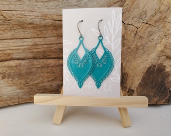 May include: Teal-colored, filigree-style earrings with a teardrop shape. The earrings are displayed on a small easel, with a card behind them that has the text "Elizabeth Q Designs". The background is a wooden surface.