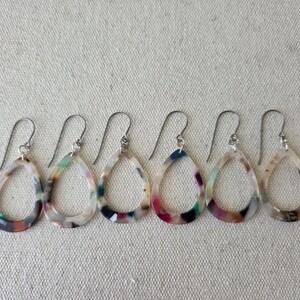 May include: A row of six teardrop-shaped earrings with silver hooks. The earrings have a marbled pattern in shades of white, brown, pink, blue, and green. The earrings are displayed on a textured, off-white surface.