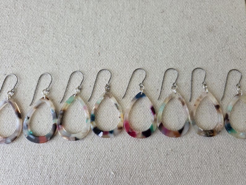 May include: A row of six teardrop-shaped earrings with silver hooks. The earrings have a marbled pattern in shades of white, brown, pink, blue, and green. The earrings are displayed on a textured, off-white surface.