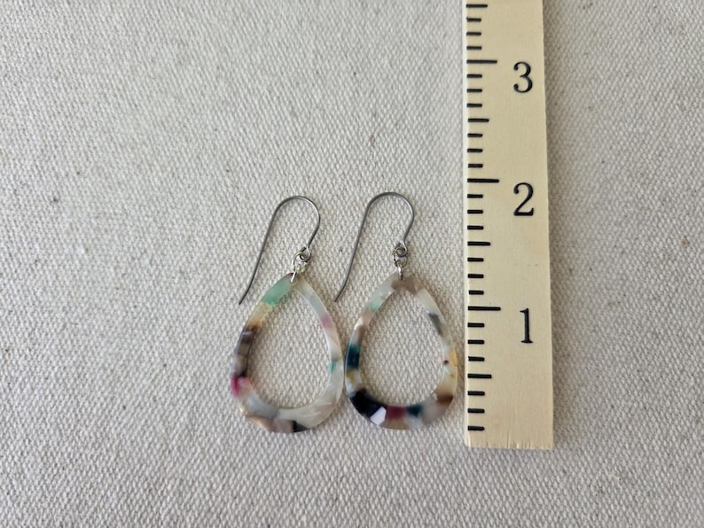 May include: A pair of teardrop earrings with a colorful, marbled pattern. The earrings are approximately 1.5 inches long and feature a silver-tone hook. The colors include white, pink, green, and black.
