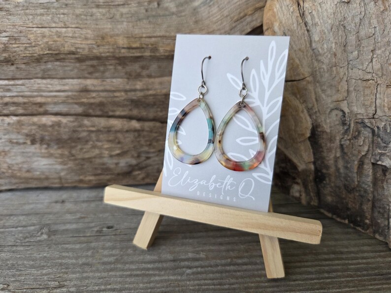 May include: A pair of teardrop-shaped earrings with a colorful, marbled pattern. The earrings are displayed on a small easel with a card that reads "Elizabeth Q Designs". The earrings have silver-colored hooks.