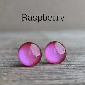 May include: A pair of round stud earrings in a vibrant raspberry pink color with a smooth, glossy finish. The word Raspberry is printed in black text above the earrings, which feature a reflective surface and a circular, dome-like shape.