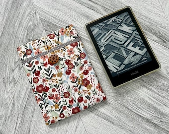 Op de afbeelding: Een witte Kindle e-reader hoes van stof met een bloemenpatroon in tinten rood, roze en geel. De hoes heeft een knoopsluiting en wordt getoond met een Kindle e-reader.